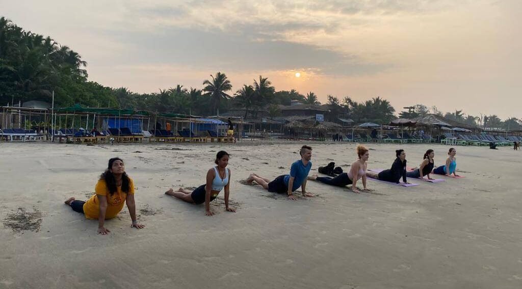 Seven people doing backbend poses in a row on a sandy beach with palm trees behind