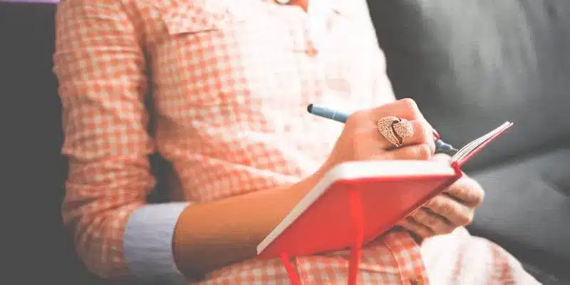 Person in a checkered shirt writing in a red journal with a pen