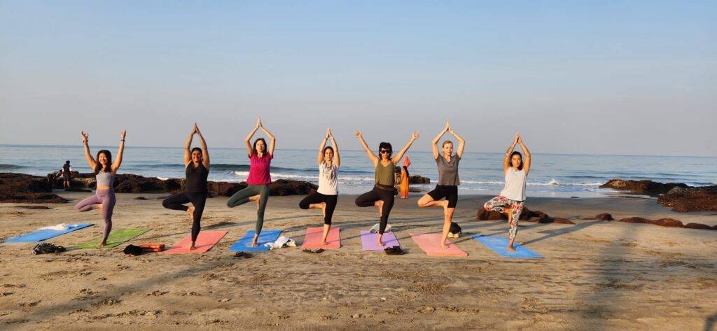 Seven people in yoga tree pose on colorful mats along the beach