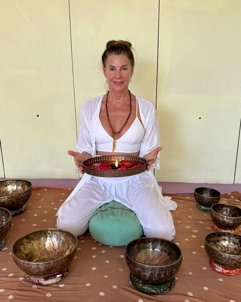 "Woman in white holding a lit lamp and rose petal tray, surrounded by brass singing bowls
