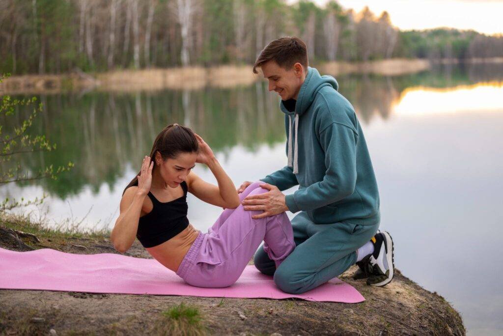Woman doing a sit-up on a pink mat by a lake while a man holds her knees