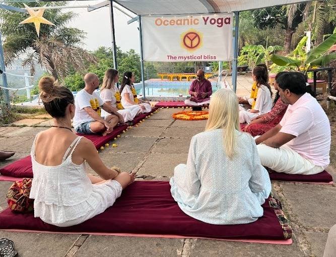 Group seated on maroon mats facing a man beside a flower arrangement under an Oceanic Yoga banner