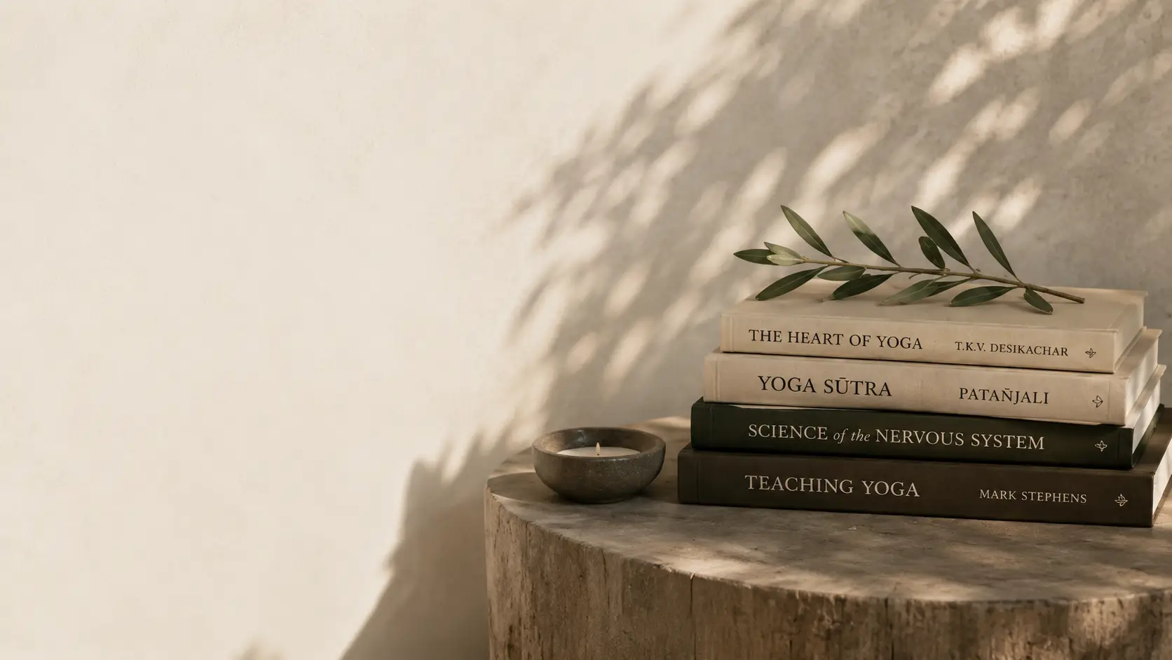 Stack of yoga books with an olive sprig beside a lit candle on a wooden table